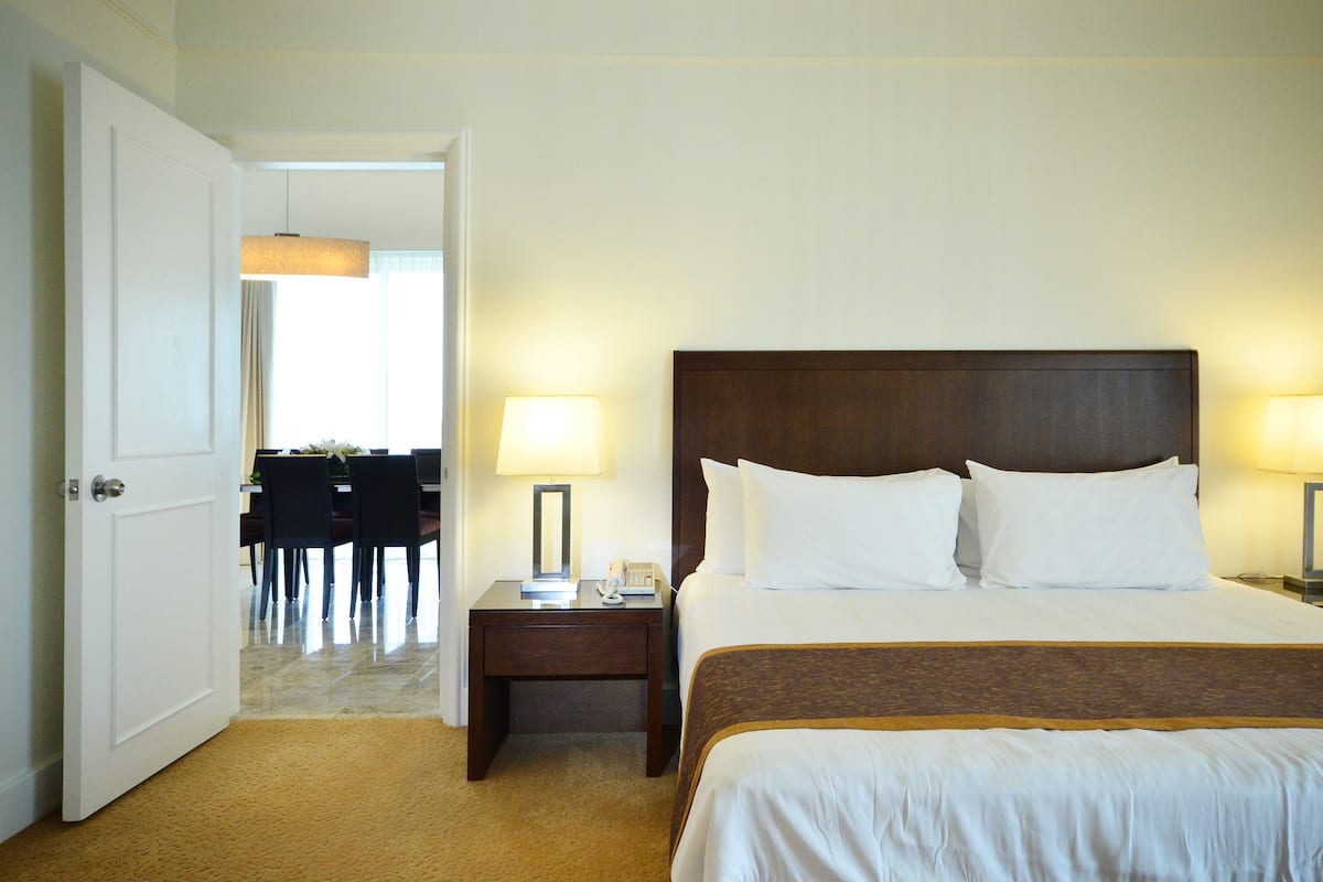 A hotel room with a bed, two bedside lamps, a nightstand, and a door leading to a dining area.