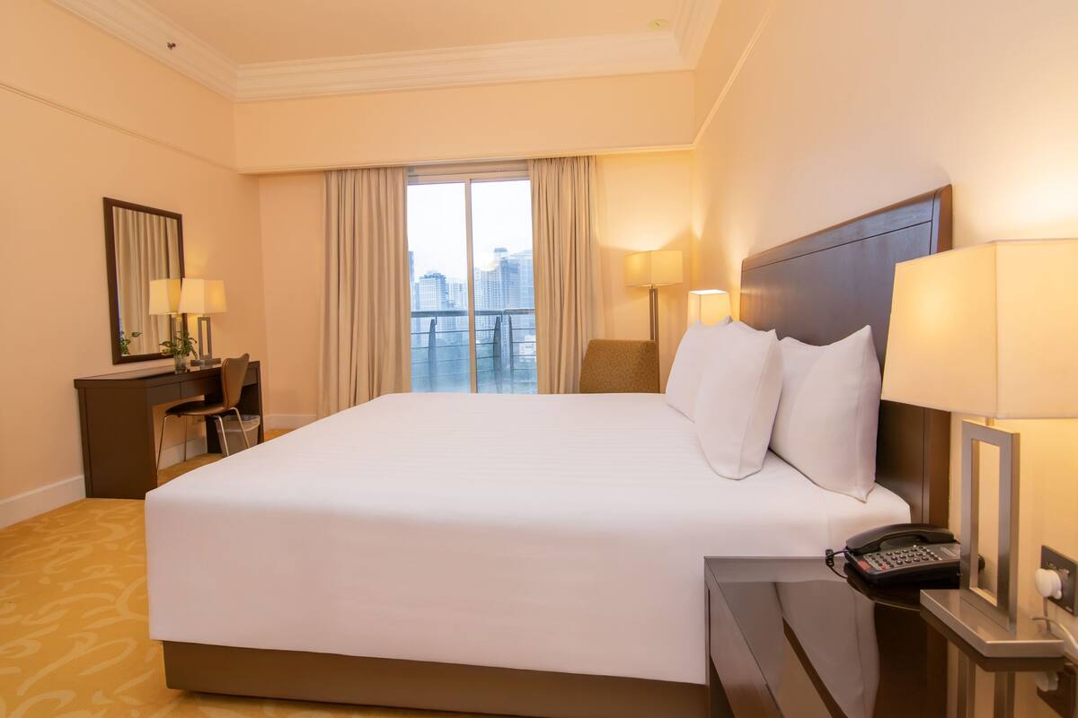 A hotel room with a large bed, a desk with a lamp, a chair, a phone, and a view of the city.