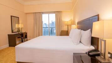 A hotel room with a large bed, a desk with a lamp, a chair, a phone, and a view of the city.