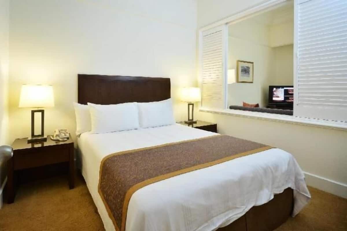 Una habitación de hotel con cama, dos mesitas de noche, un tocador, un espejo y una ventana con persianas.