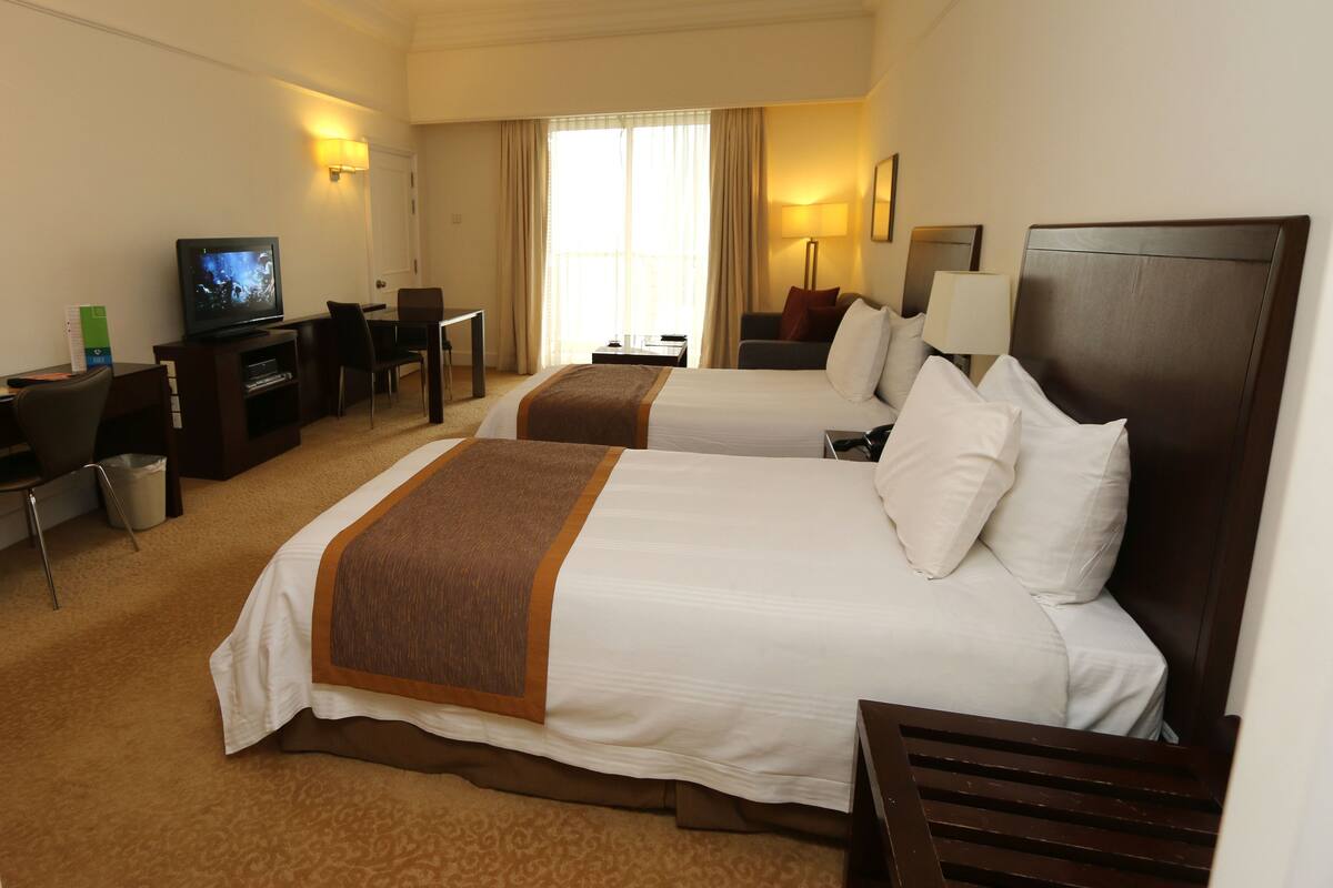 A hotel room with two beds, a desk, a television, and a window with curtains.