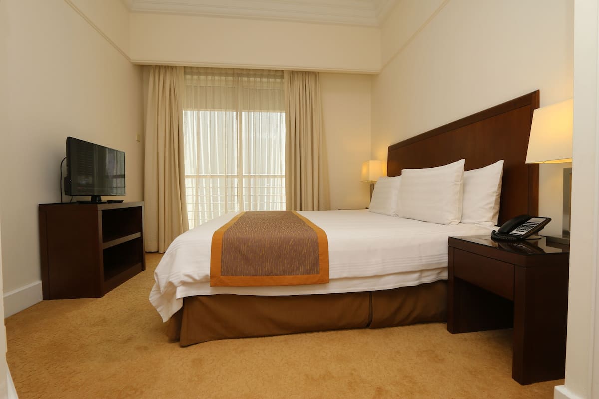 A hotel room with a bed, a TV on a stand, a bedside table with a lamp, and a window with curtains.