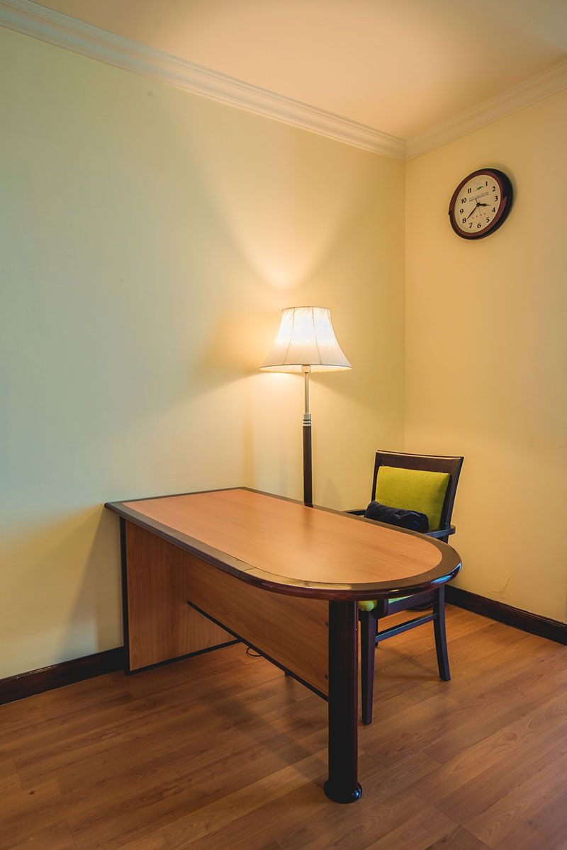 A room with a wooden table, a lamp, a chair, and a wall clock.