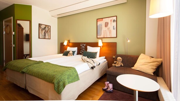 A hotel room with a double bed, a sofa, a small table, and a teddy bear.