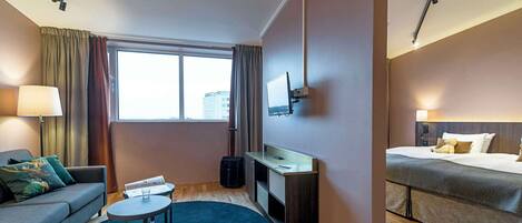 A hotel room with a sofa, a desk, a bed, a TV mounted on the wall, and a large window.