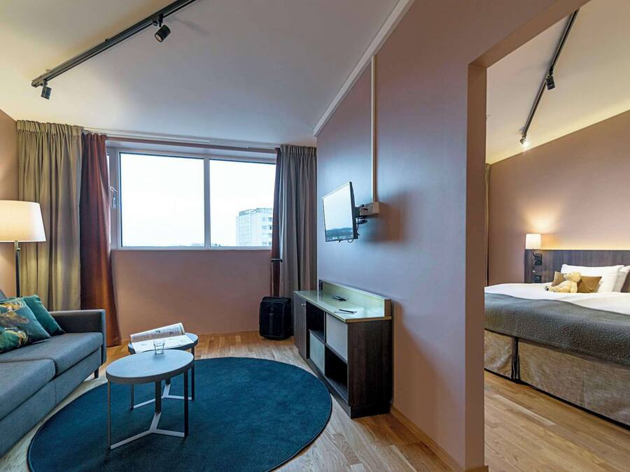 Ett hotellrum med en soffa, ett skrivbord, en säng, en tv som är monterad på väggen och ett stort fönster.