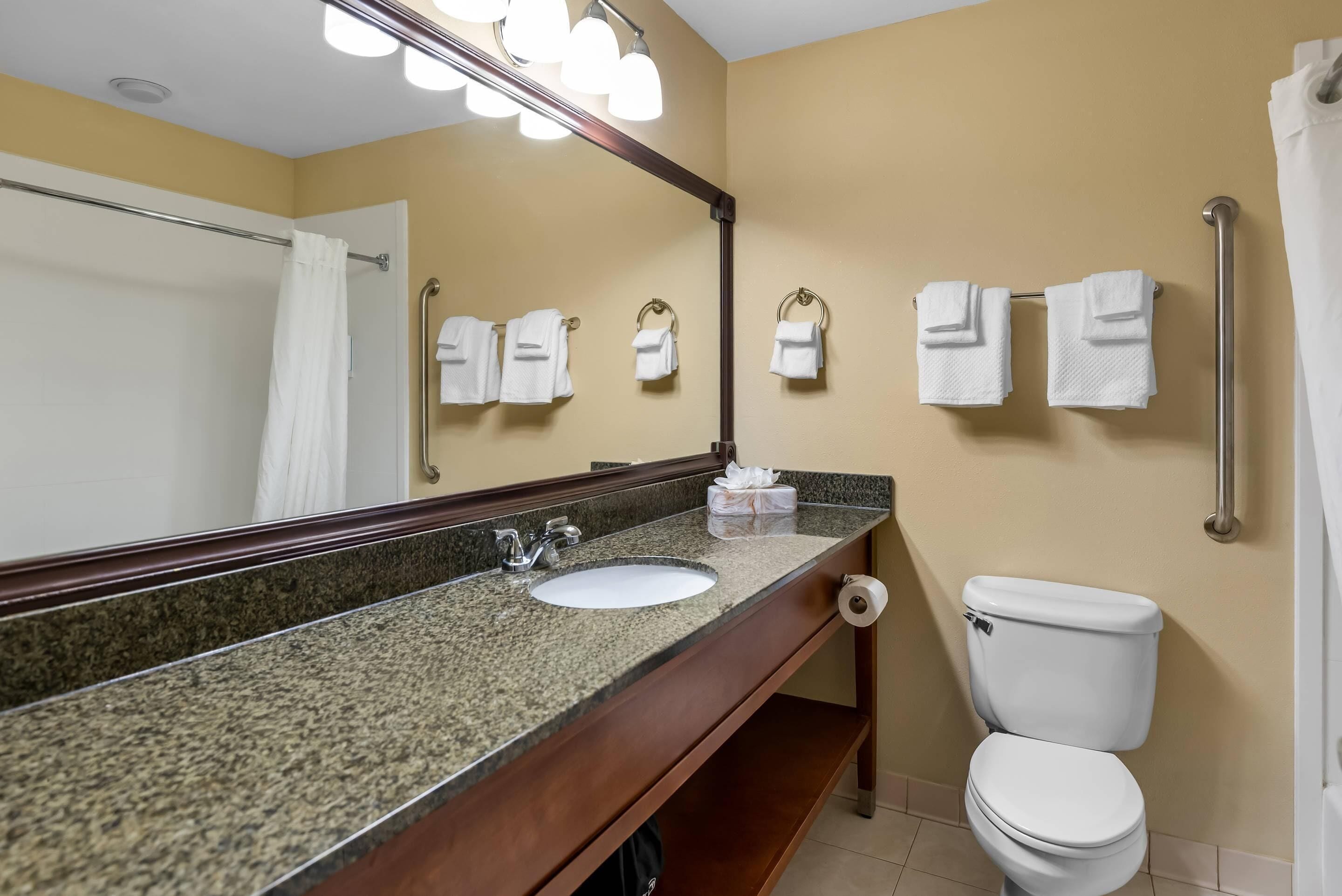 A bathroom with a granite countertop sink, a mirror, a white toilet, and a towel rack with multiple towels.