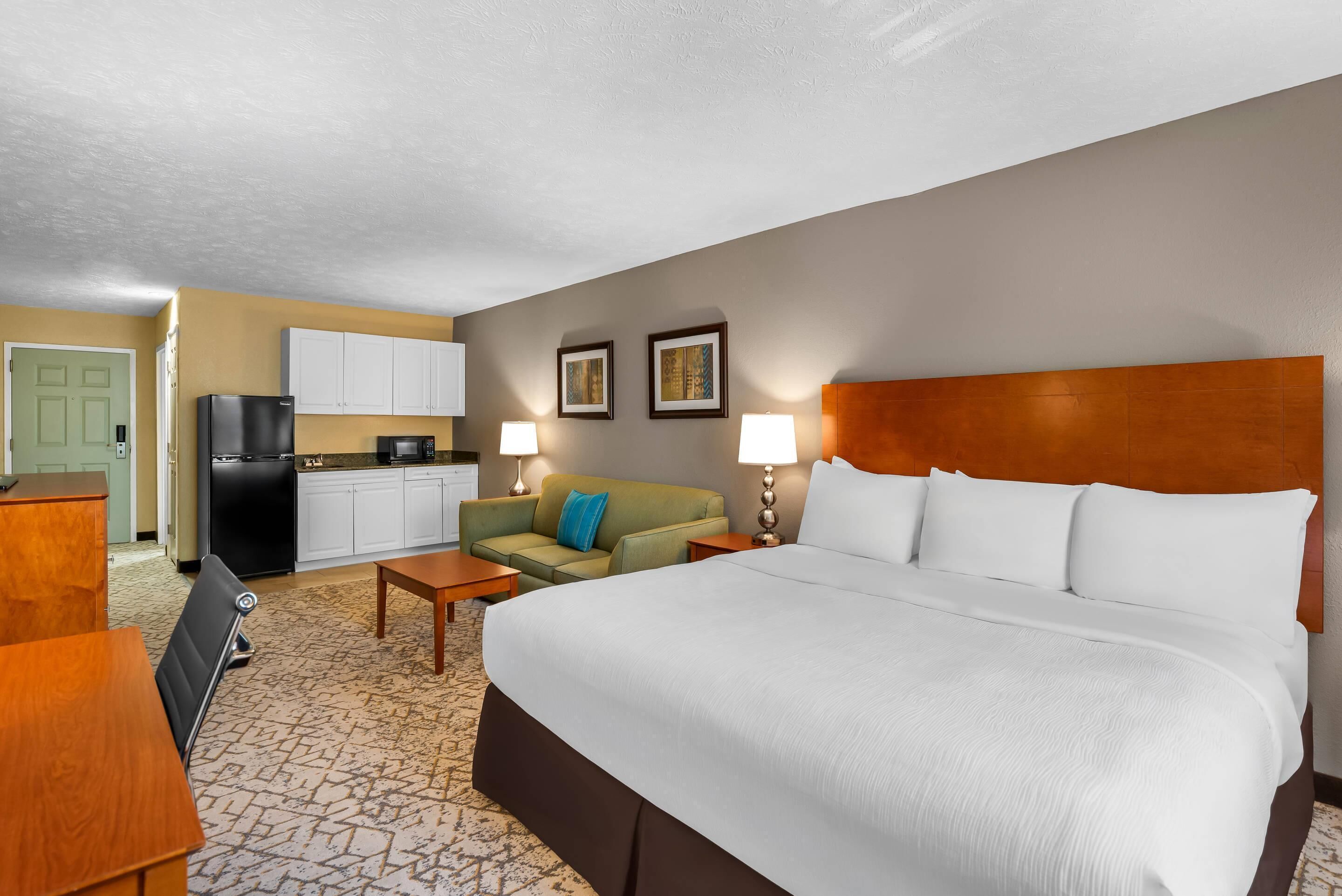 A hotel room with a bed, a sofa, a small table, a kitchen area, and a door.
