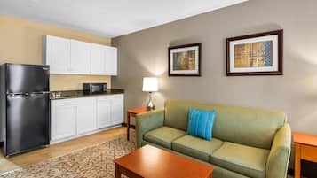 A compact living space with a kitchenette, a green sofa with a blue pillow, a wooden coffee table, and framed artwork on the wall.