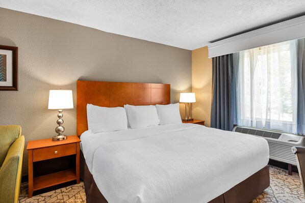 A hotel room with a large bed, two bedside tables with lamps, a chair, a framed picture on the wall, and a window with curtains.