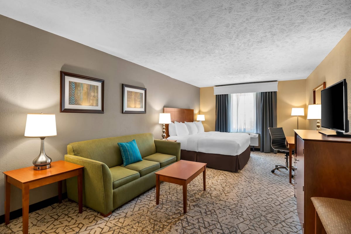 A hotel room with a bed, a green sofa, a wooden side table, a small wooden table, a TV, and two wall-mounted lamps.