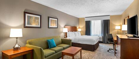 A hotel room with a bed, a green sofa, a wooden side table, a small wooden table, a TV, and two wall-mounted lamps.