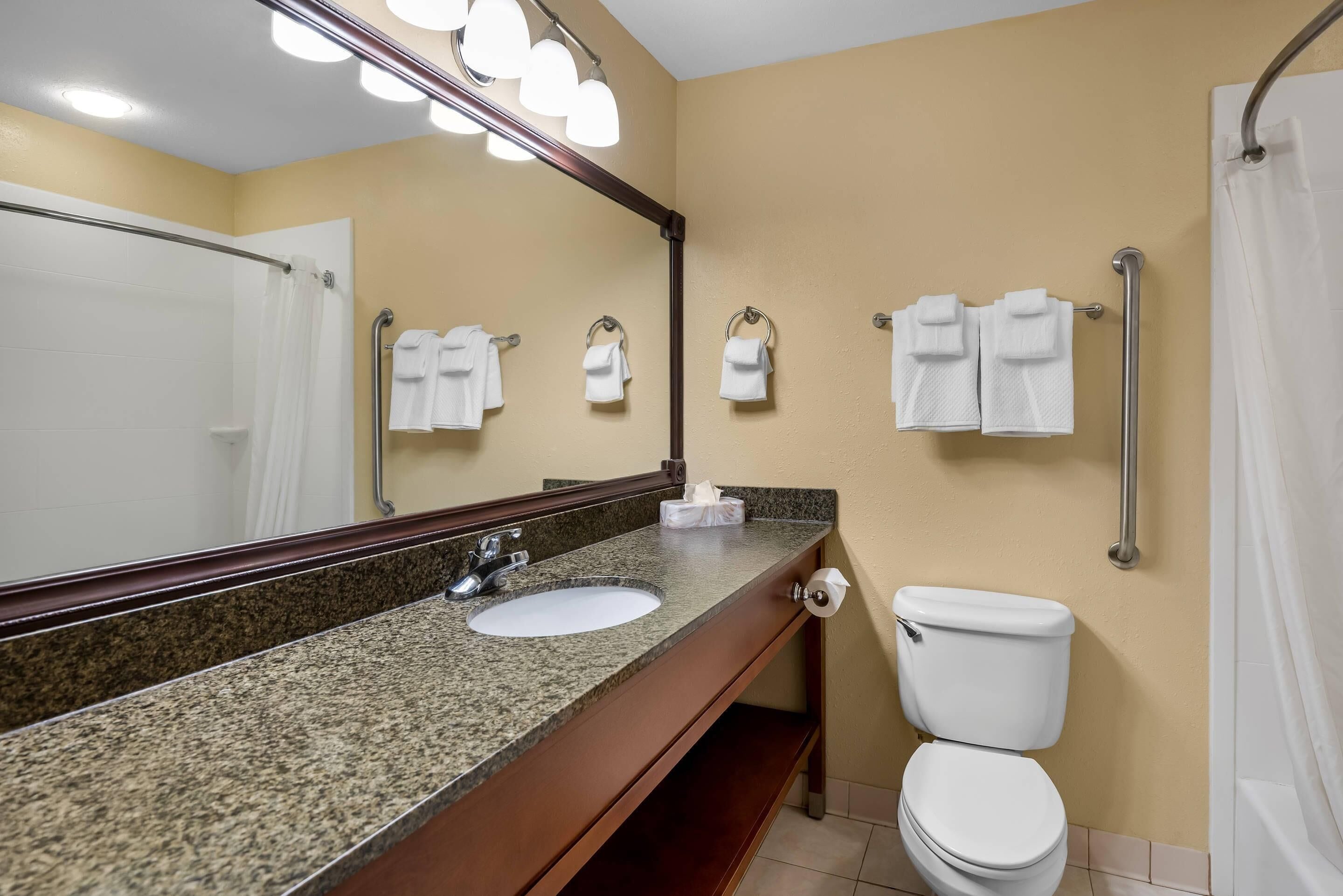A bathroom with a granite countertop sink, a mirror, a toilet, and multiple towels hanging on a rack.