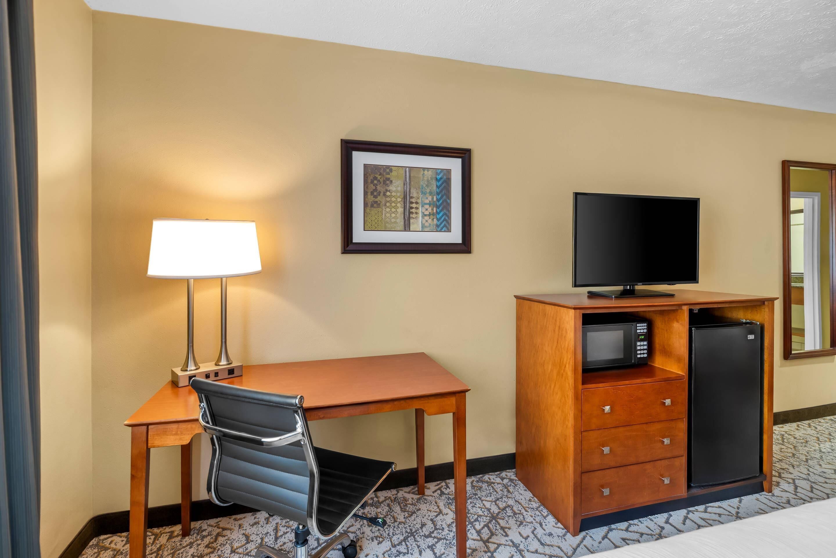A hotel room with a desk, chair, lamp, television, microwave, refrigerator, and a framed picture on the wall.