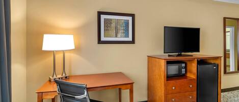 A hotel room with a desk, chair, lamp, television, microwave, refrigerator, and a framed picture on the wall.