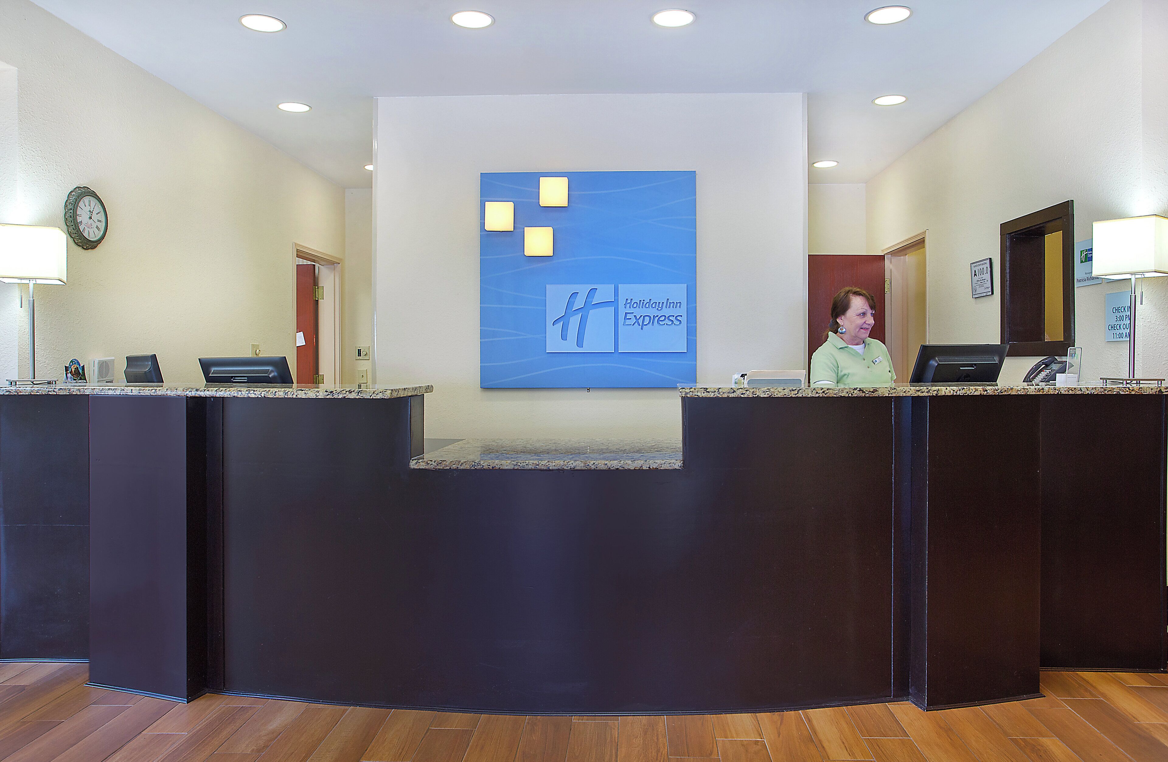 A hotel reception desk with a clock on the wall, a sign for Holiday Inn Express, and a reception desk with a computer and phone.
