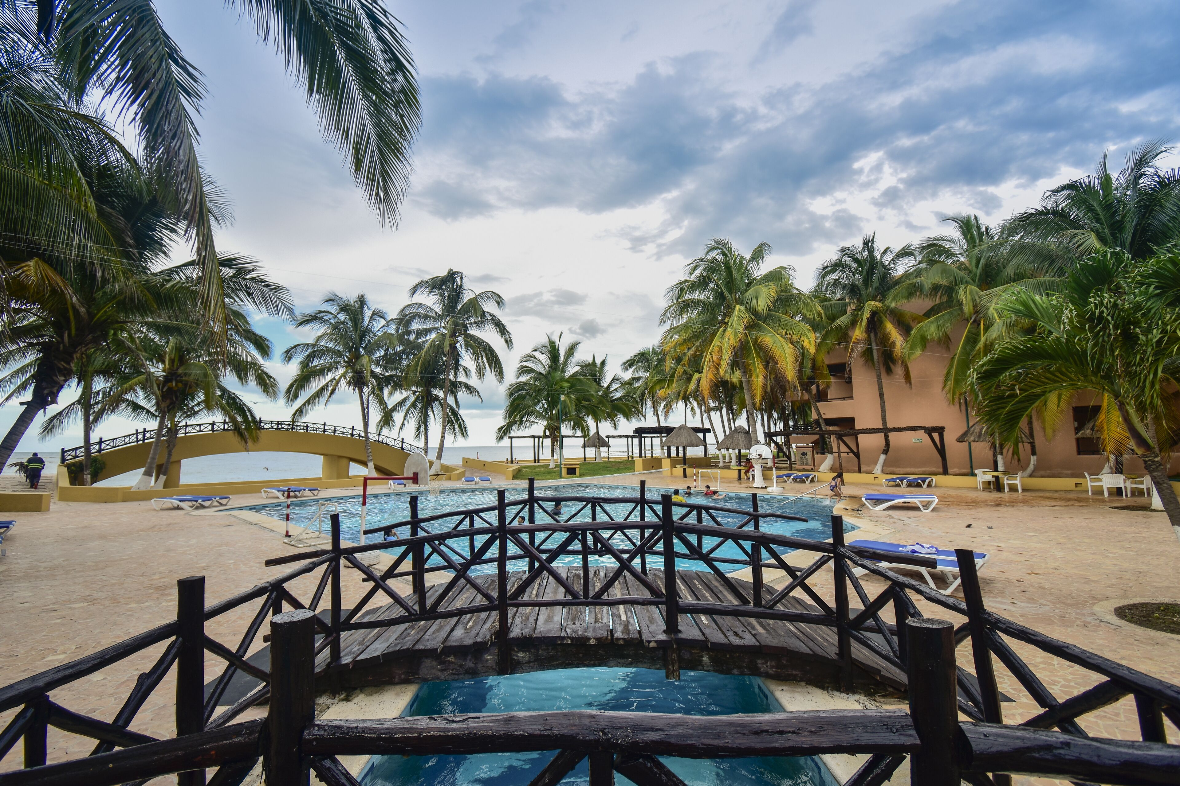 A pool area with a wooden deck, palm trees, and a bridge.