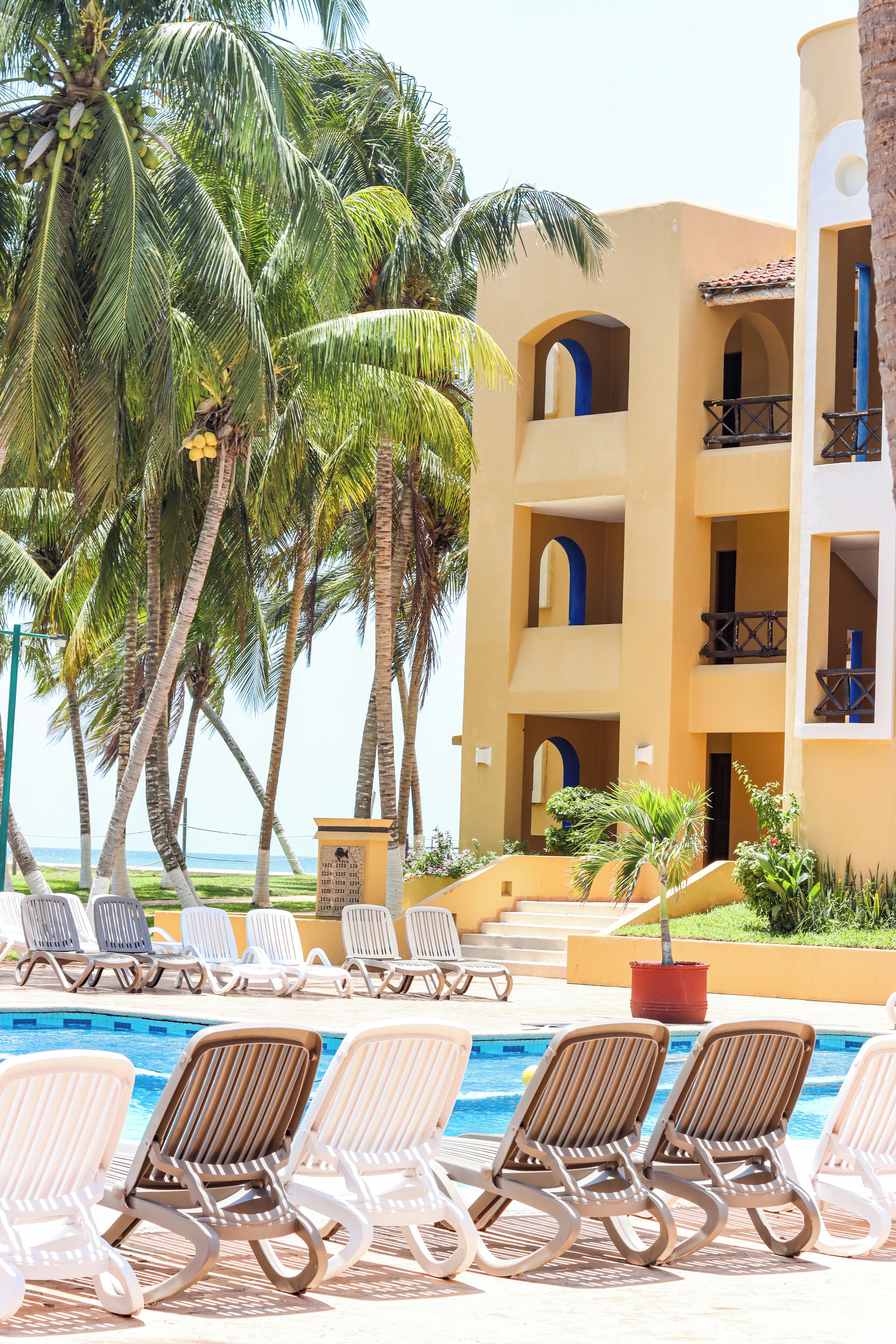 Un espace aménagé au bord de la piscine, avec des chaises longues, des palmiers et un bâtiment doté de balcons.