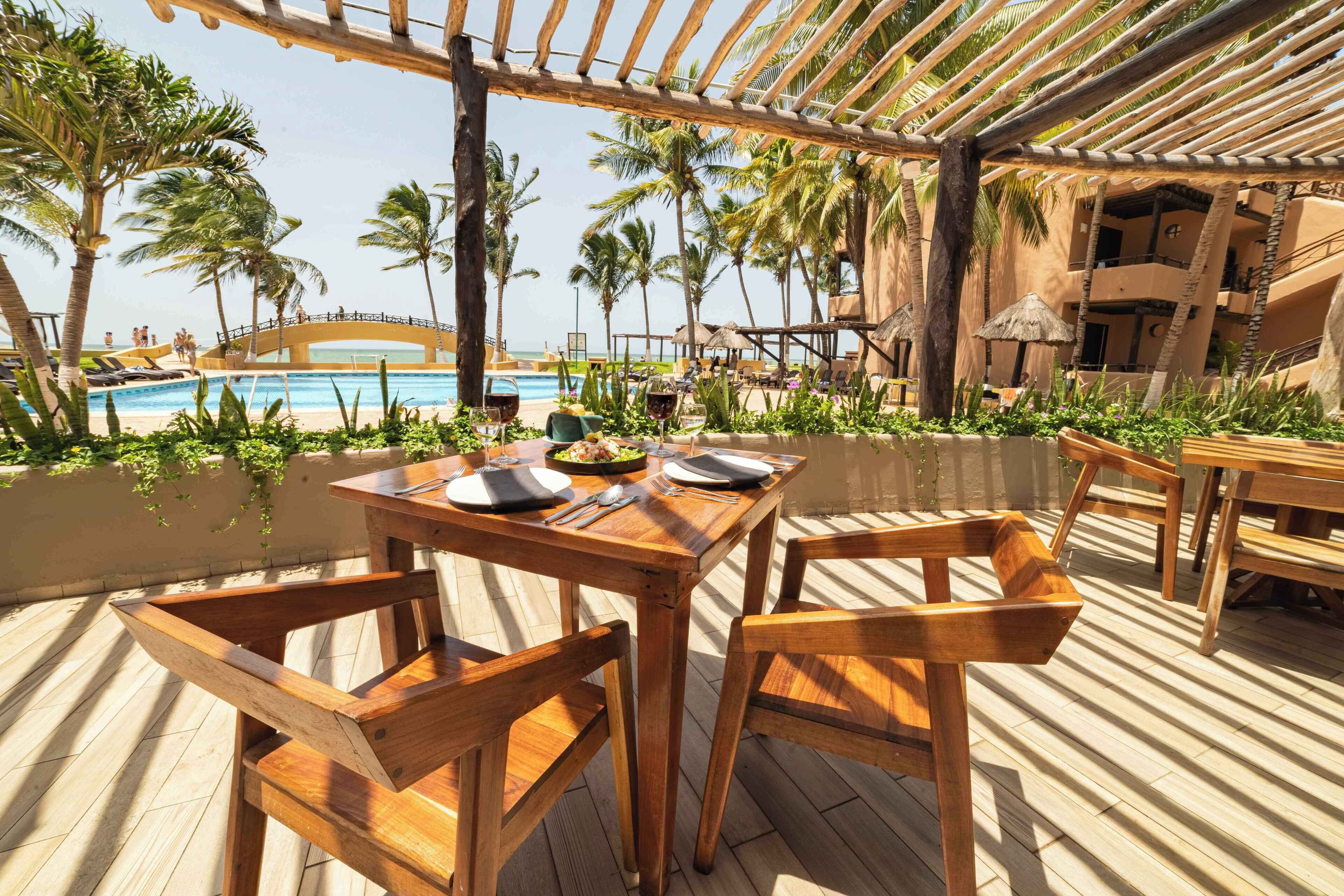 An outdoor dining area with wooden tables and chairs, overlooking a pool and palm trees.