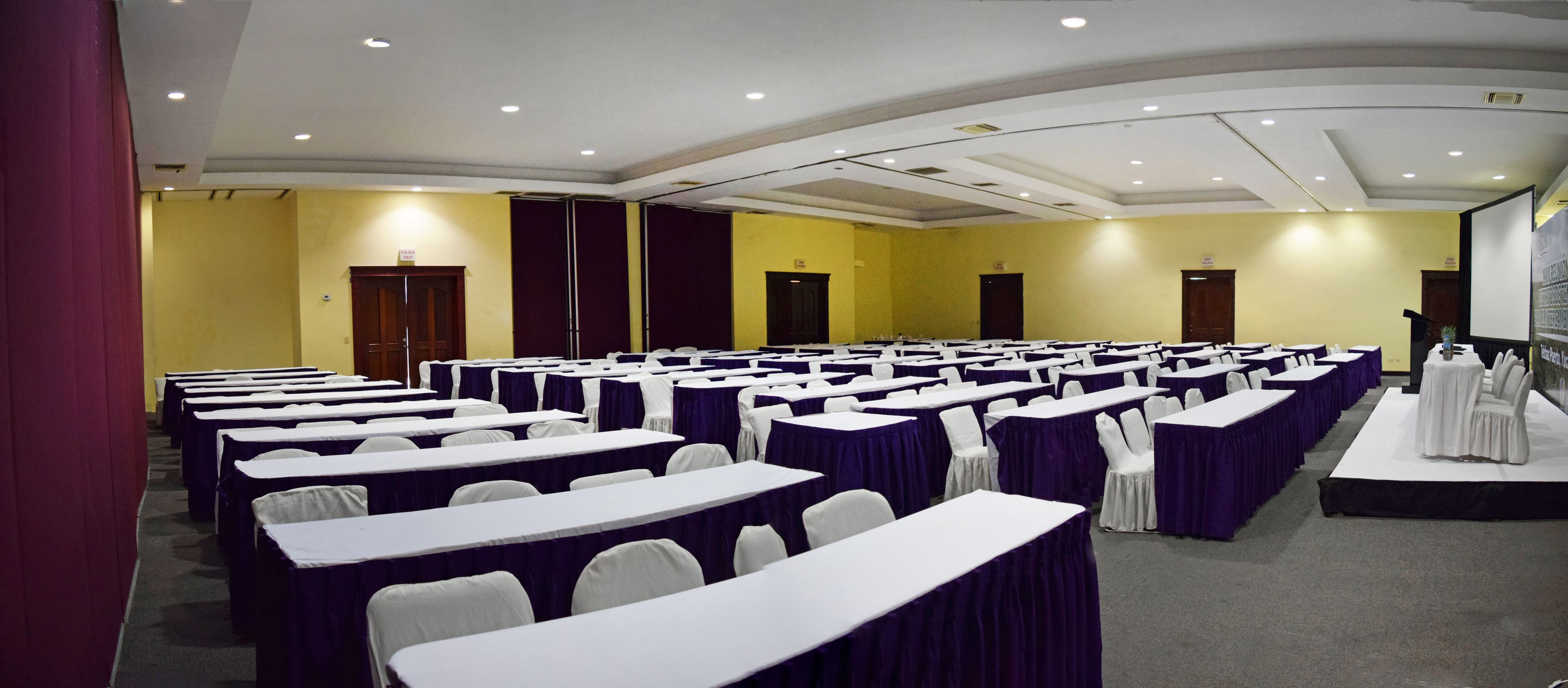 A conference room with rows of tables and chairs, a stage, and a yellow wall.