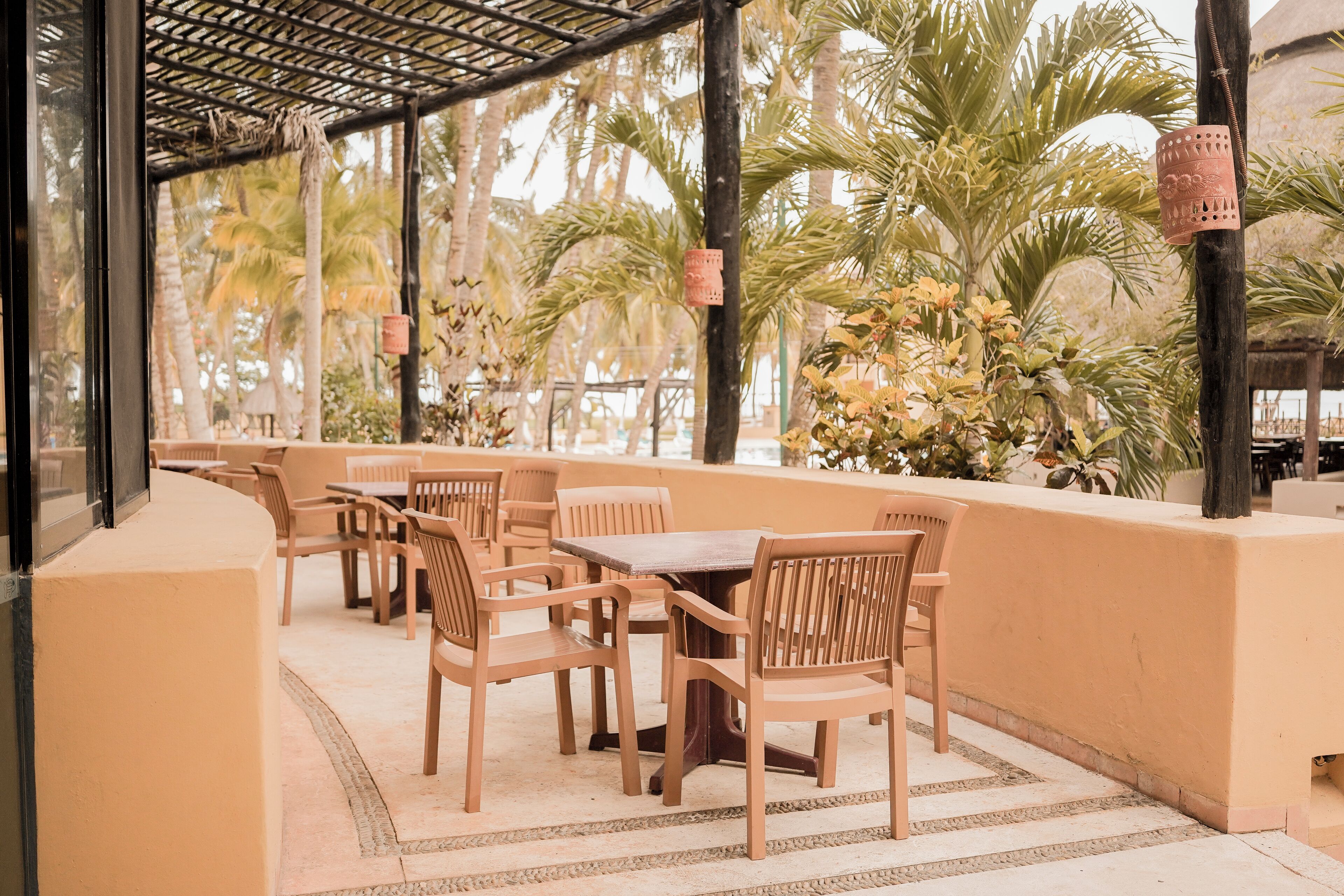 An outdoor seating area with wooden tables and chairs, surrounded by palm trees and a thatched roof structure.