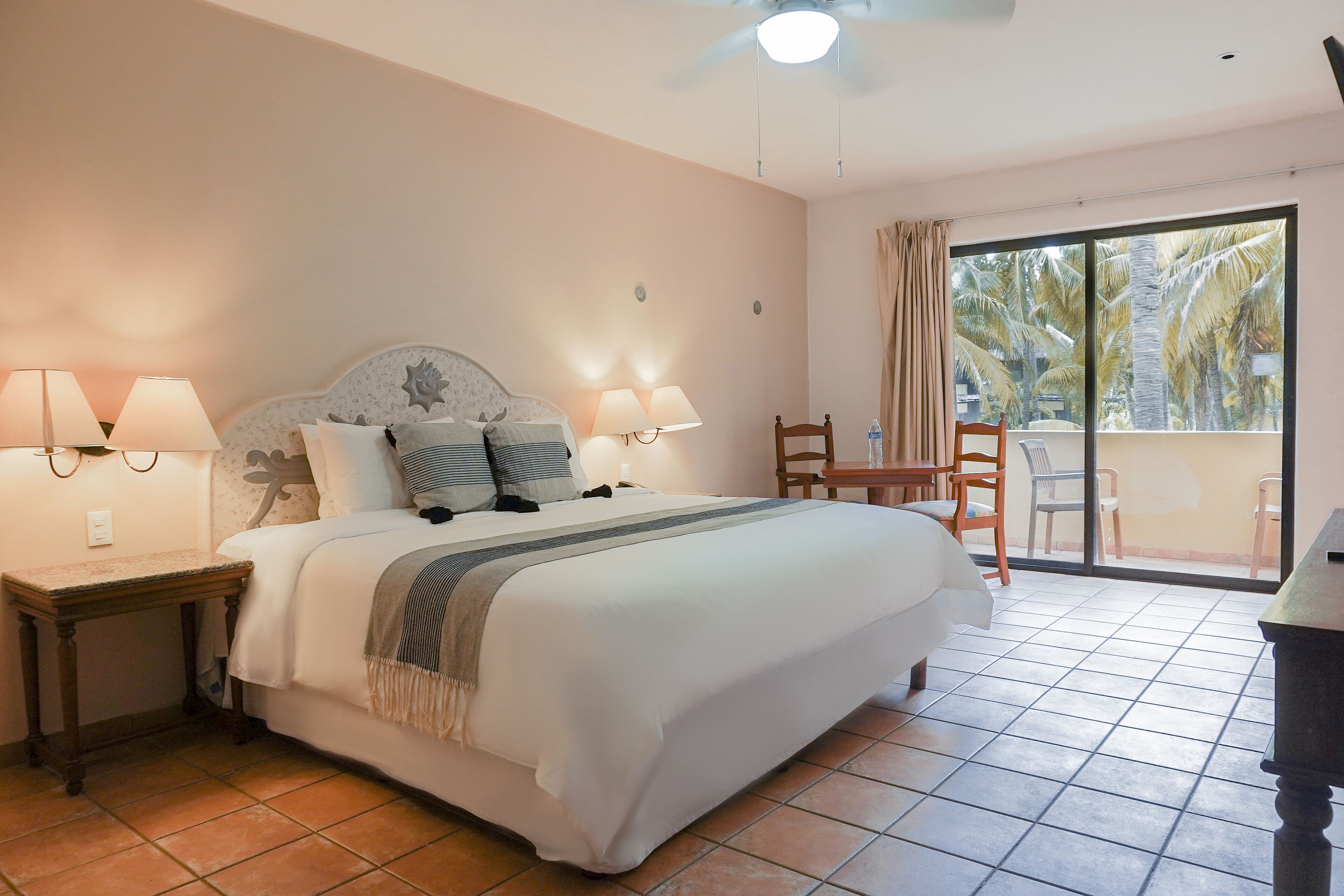 A bedroom with a bed, two bedside lamps, a nightstand, a chair, a table, and a view of palm trees through a large window.