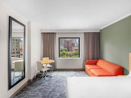 Una habitación de hotel con cama, sofá, mesa de comedor con sillas, ventana grande con vista a la ciudad y espejo.