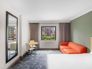 A hotel room with a bed, a sofa, a dining table with chairs, a large window with a city view, and a mirror. - Mercure Melbourne Southbank (Southbank)