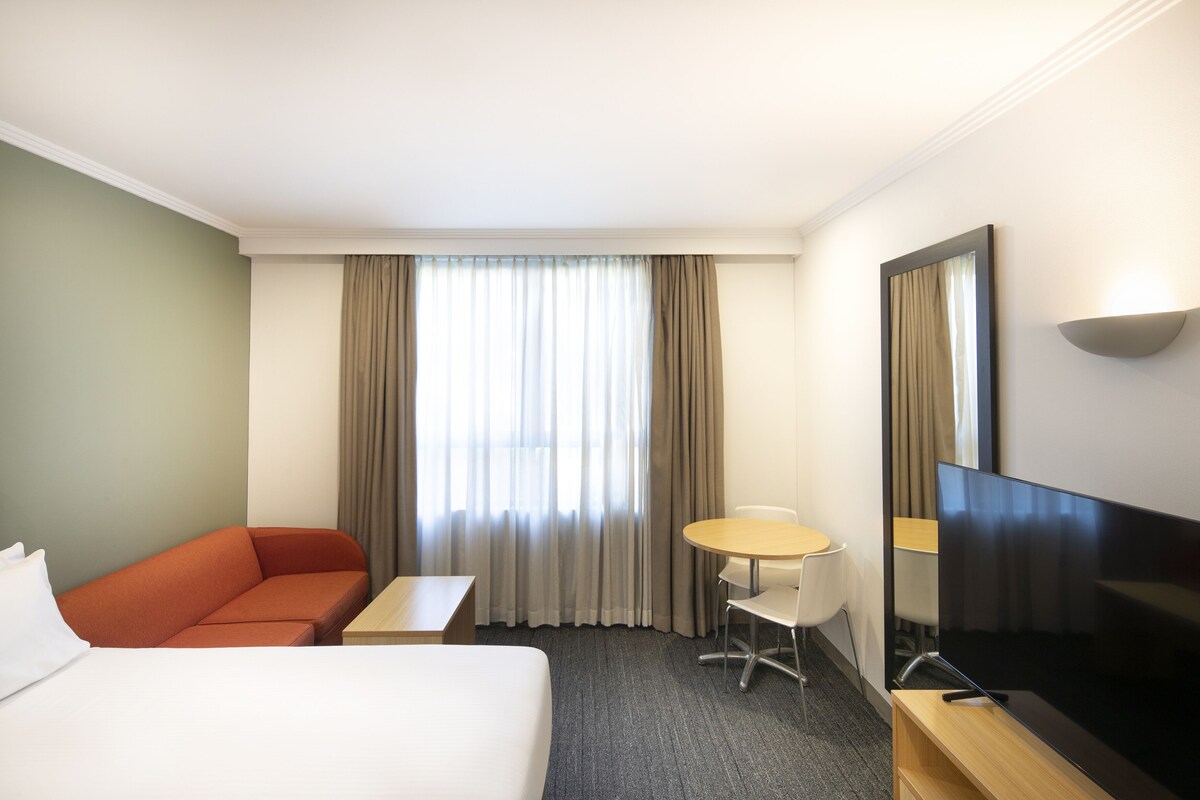 Une chambre d’hôtel avec un lit, un canapé, une petite table, une chaise, une télévision et une fenêtre avec des rideaux.