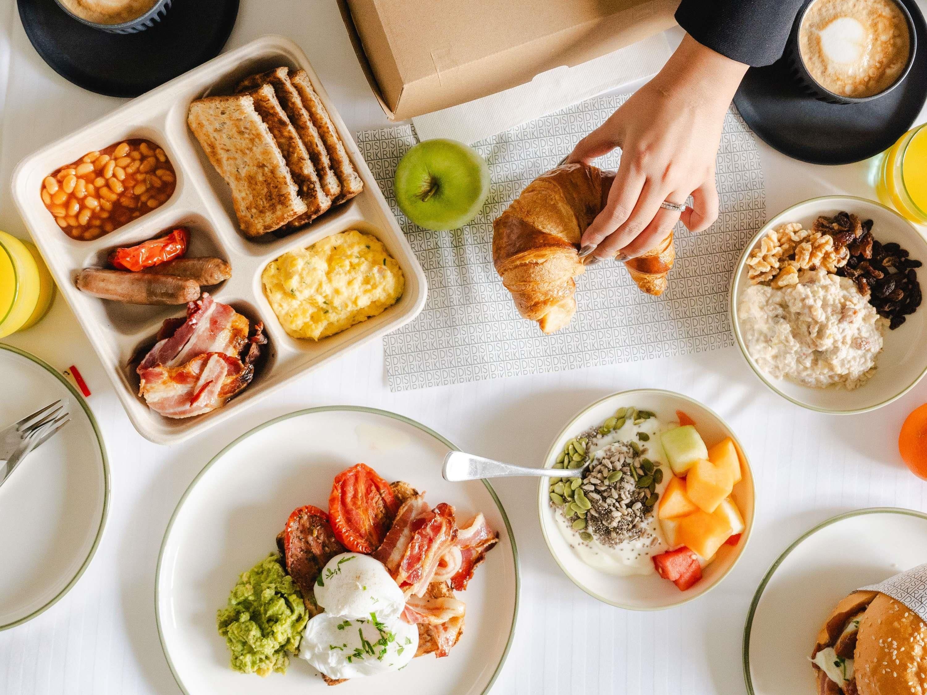 Una mesa con una variedad de alimentos para el desayuno, incluyendo tostadas, frijoles, salchichas, huevos y un croissant.