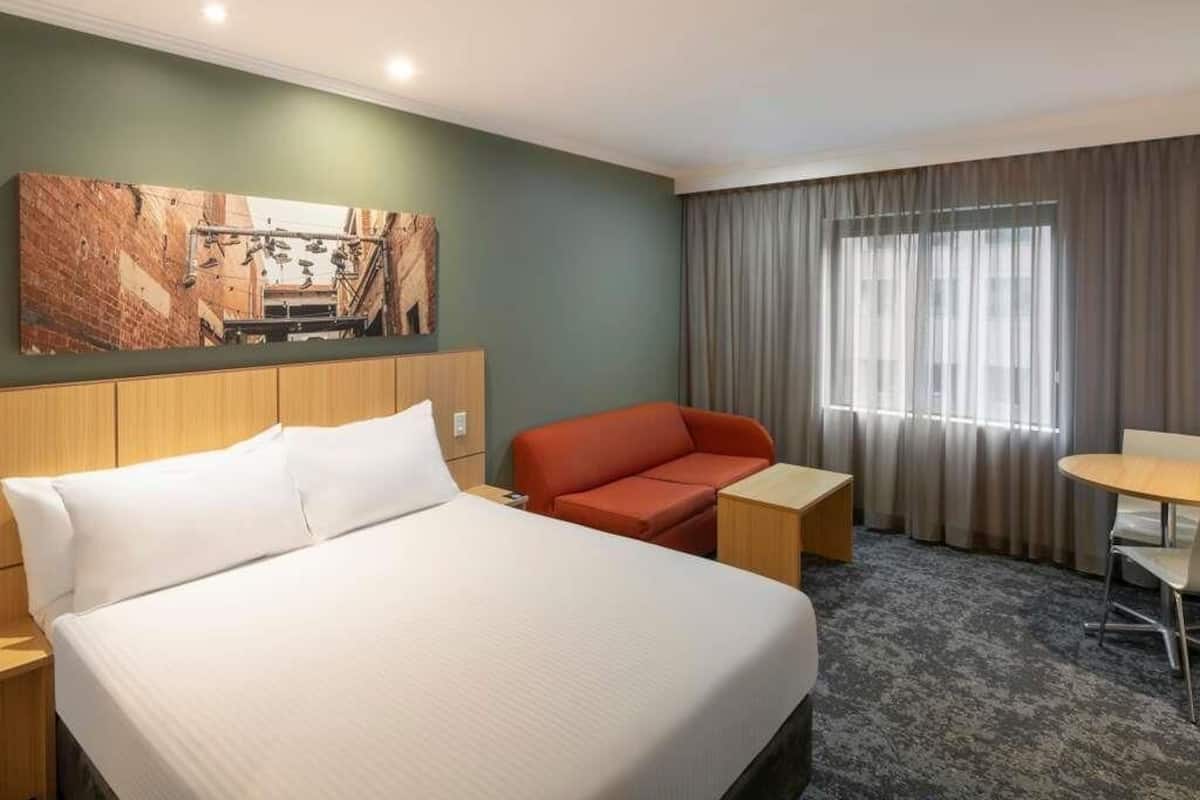 Une chambre d’hôtel avec un lit, un canapé rouge, une petite table, une chaise et un tableau au mur.