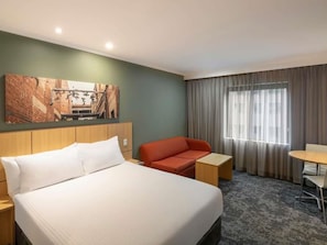 Une chambre d’hôtel avec un lit, un canapé rouge, une petite table, une chaise et un tableau au mur.