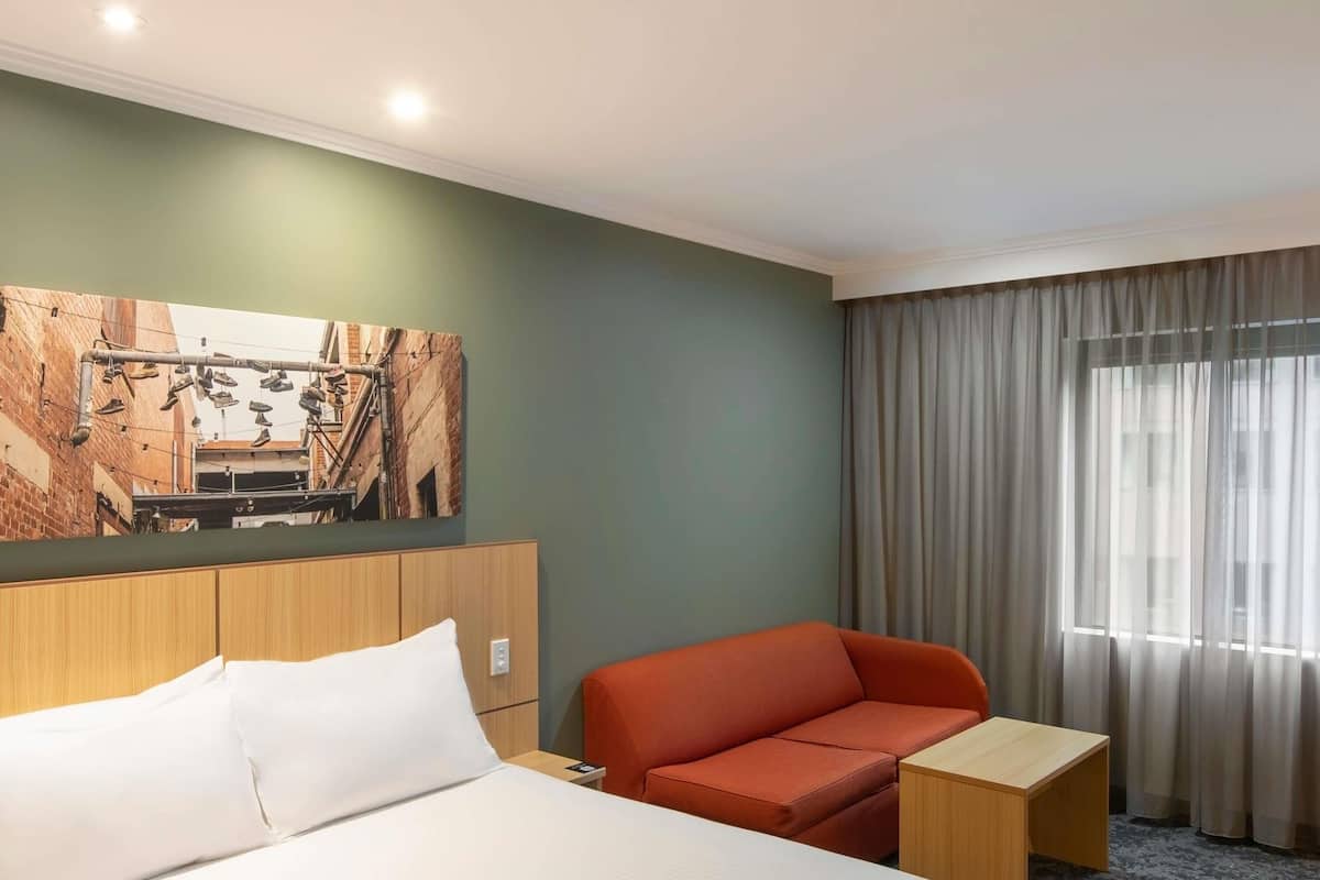 Une chambre d’hôtel avec un lit, une tête de lit en bois, un canapé rouge, une petite table en bois et une grande fenêtre avec des rideaux.