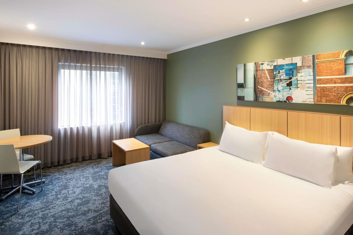 Une chambre d’hôtel comprenant un lit, un bureau, une chaise, une fenêtre avec des rideaux et un mur orné d’une fresque représentant un paysage urbain.