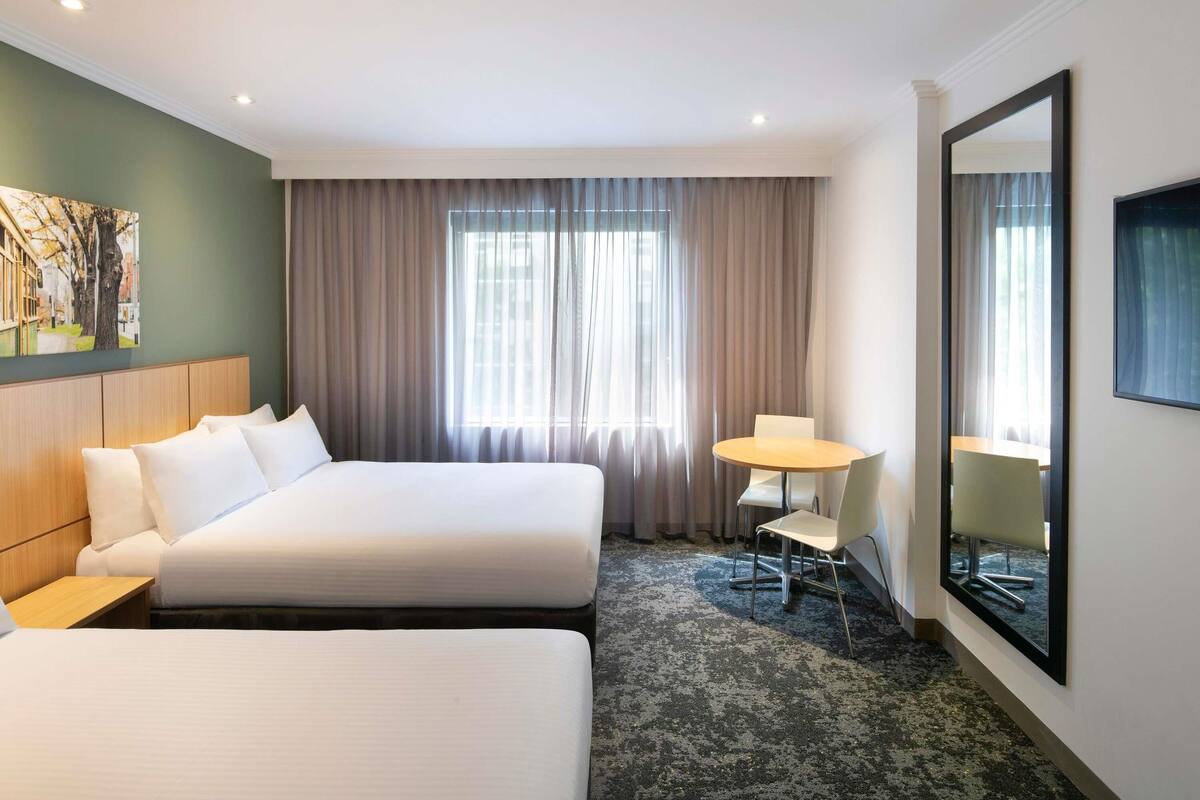 Une chambre d’hôtel avec deux lits, un bureau, une chaise, un grand miroir et une fenêtre avec des rideaux.