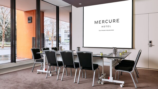 A conference room with a large screen displaying "Mercure Hotel Southbank Melbourne", six chairs, a table with a projector, and a view of the outside.