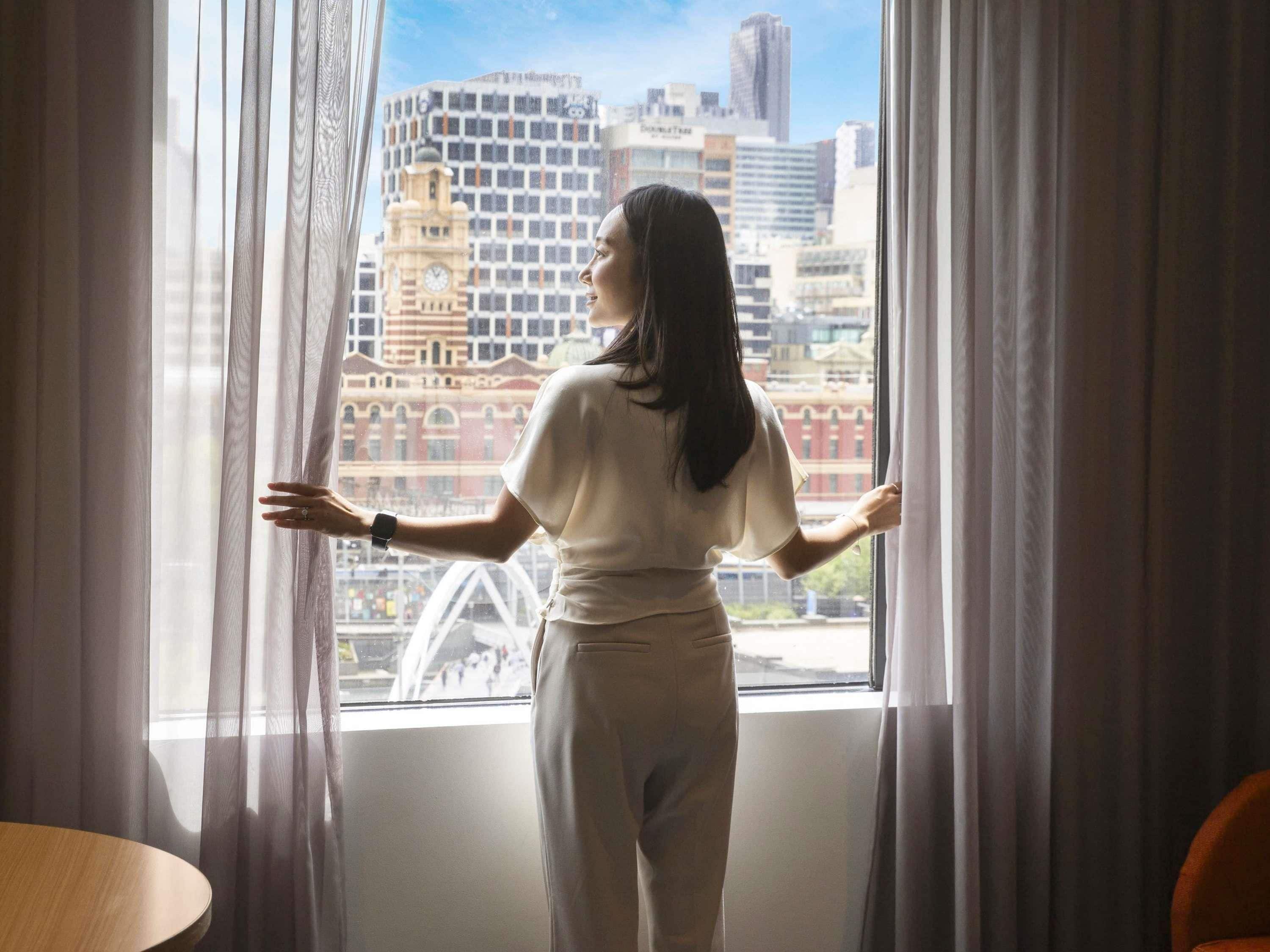 A person standing by a window with a cityscape view, curtains, and a table.