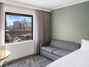A hotel room with a large window, a grey sofa, a bed, and a small table.
