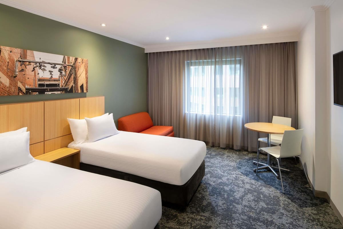 Une chambre d’hôtel avec deux lits, une petite table, une chaise, un téléviseur et une fenêtre avec des rideaux.