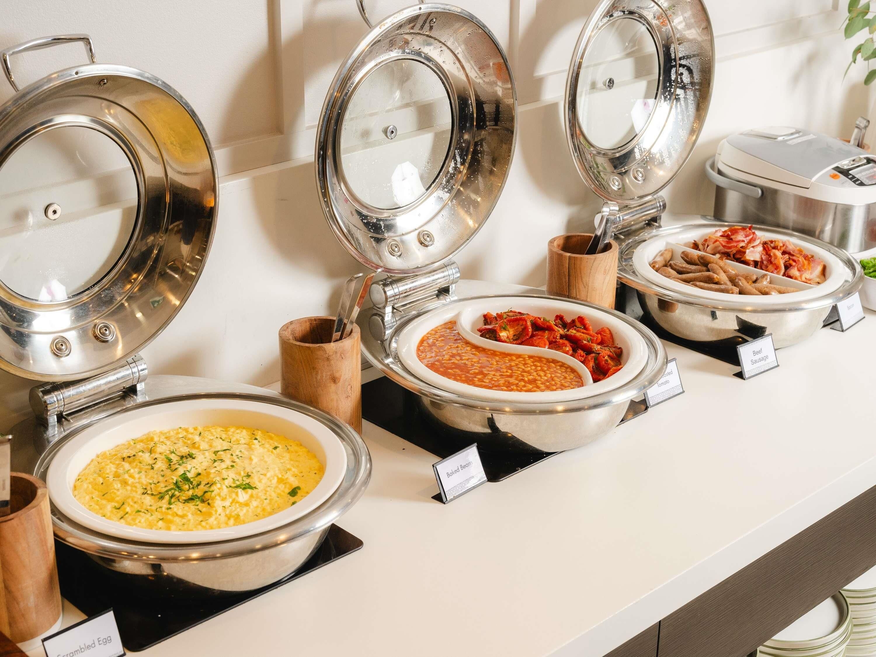 Buffet com pratos identificados, incluindo ovos mexidos, feijão assado, sopa de tomate e linguiça.