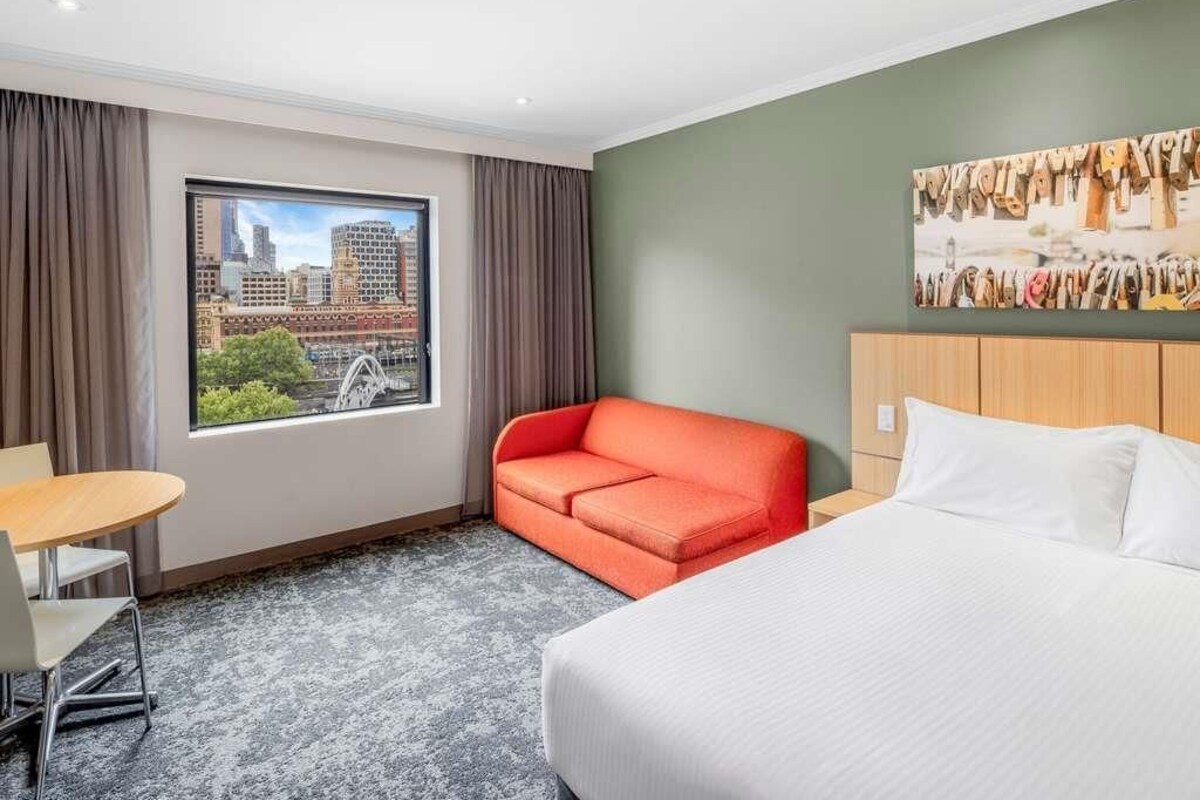 Une chambre d’hôtel avec un lit, un canapé rouge, une petite table et une grande fenêtre donnant sur le paysage urbain.
