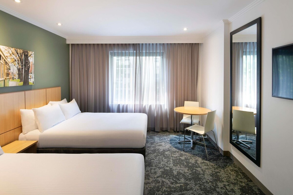 A hotel room with two beds, a desk, a chair, a large mirror, and a window with curtains.