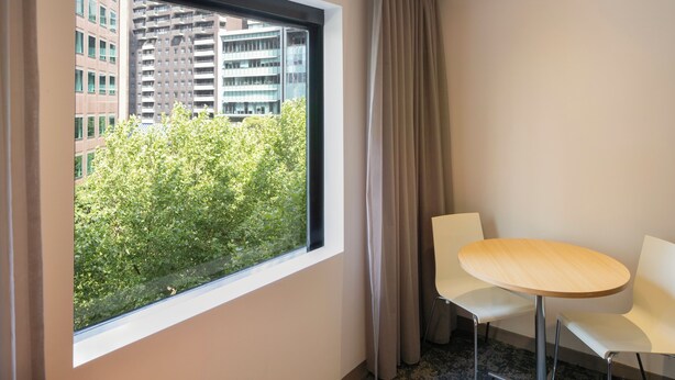 Habitación de hotel con un ventanal grande, una mesa redonda, dos sillas blancas y vista a un edificio y árboles.