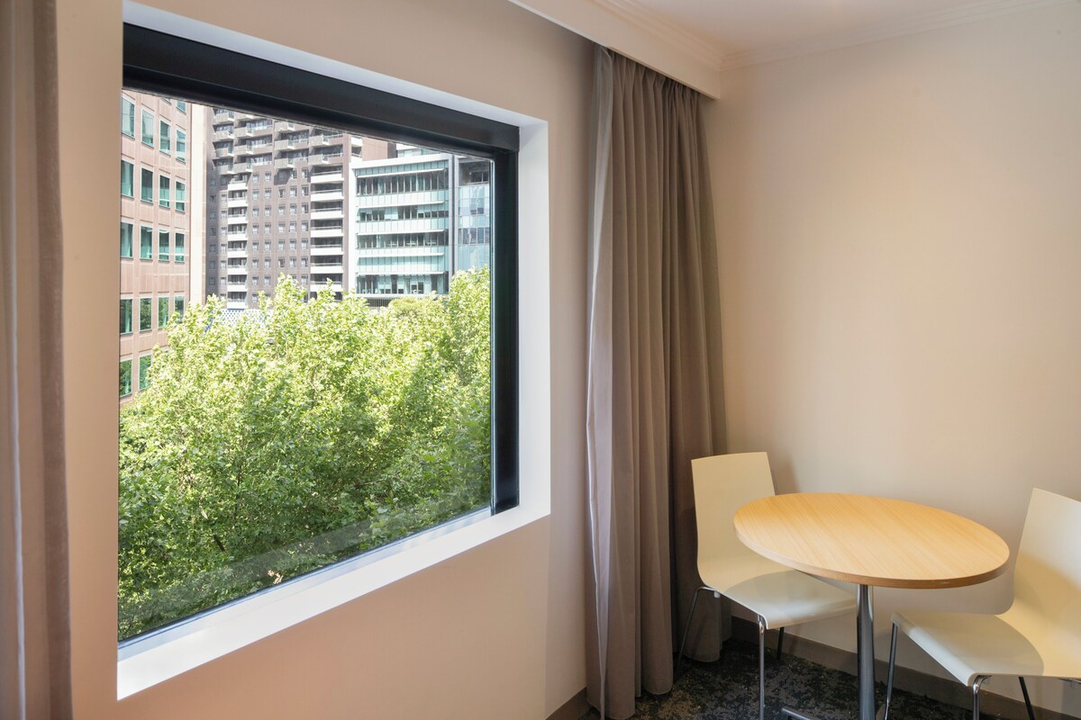 Une chambre d’hôtel avec une grande fenêtre, une table ronde, deux chaises blanches et une vue sur un immeuble et des arbres.