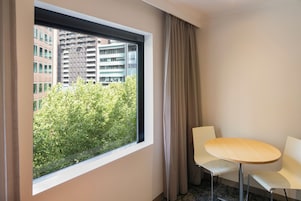 Habitación de hotel con un ventanal grande, una mesa redonda, dos sillas blancas y vista a un edificio y árboles.