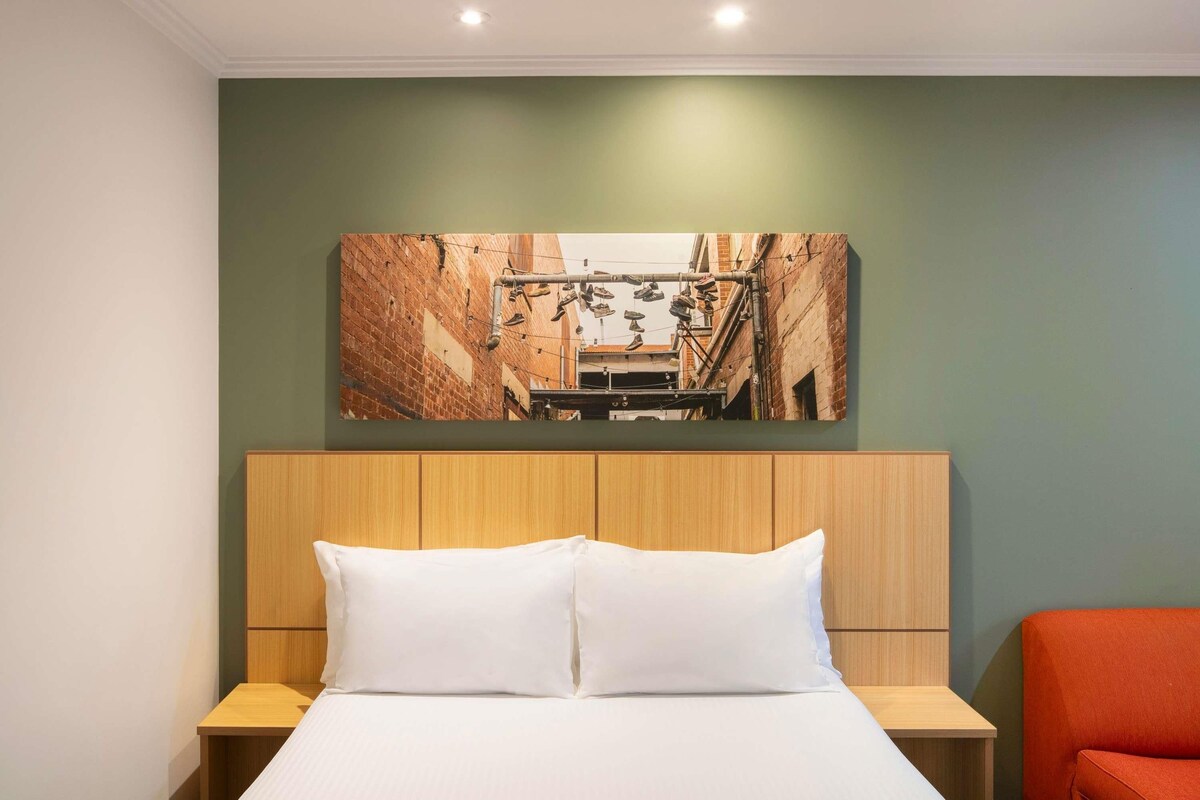 A hotel room with a bed, two nightstands, a red chair, and a large picture on the wall.