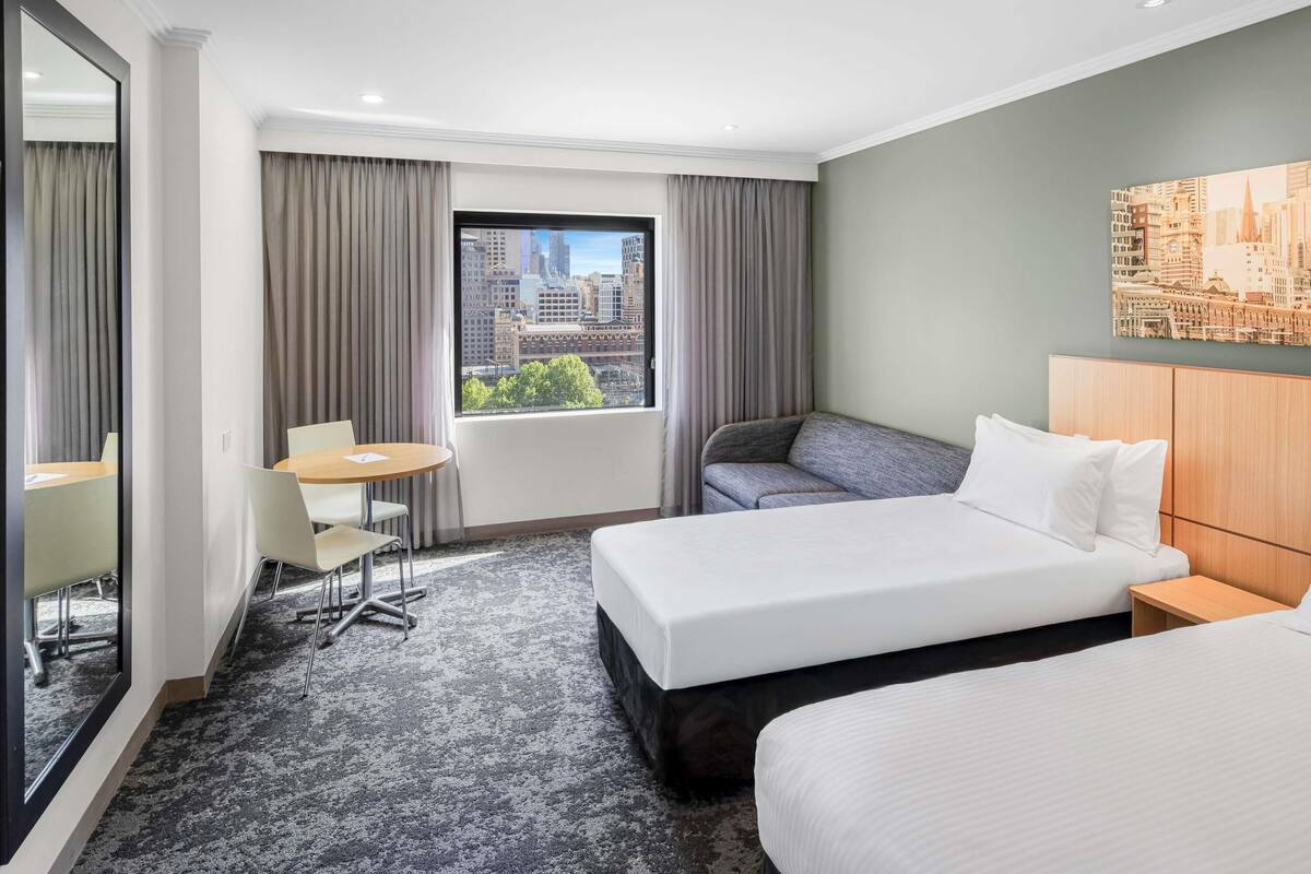 Une chambre d’hôtel avec deux lits, un bureau, une chaise et une grande fenêtre donnant sur la ville.
