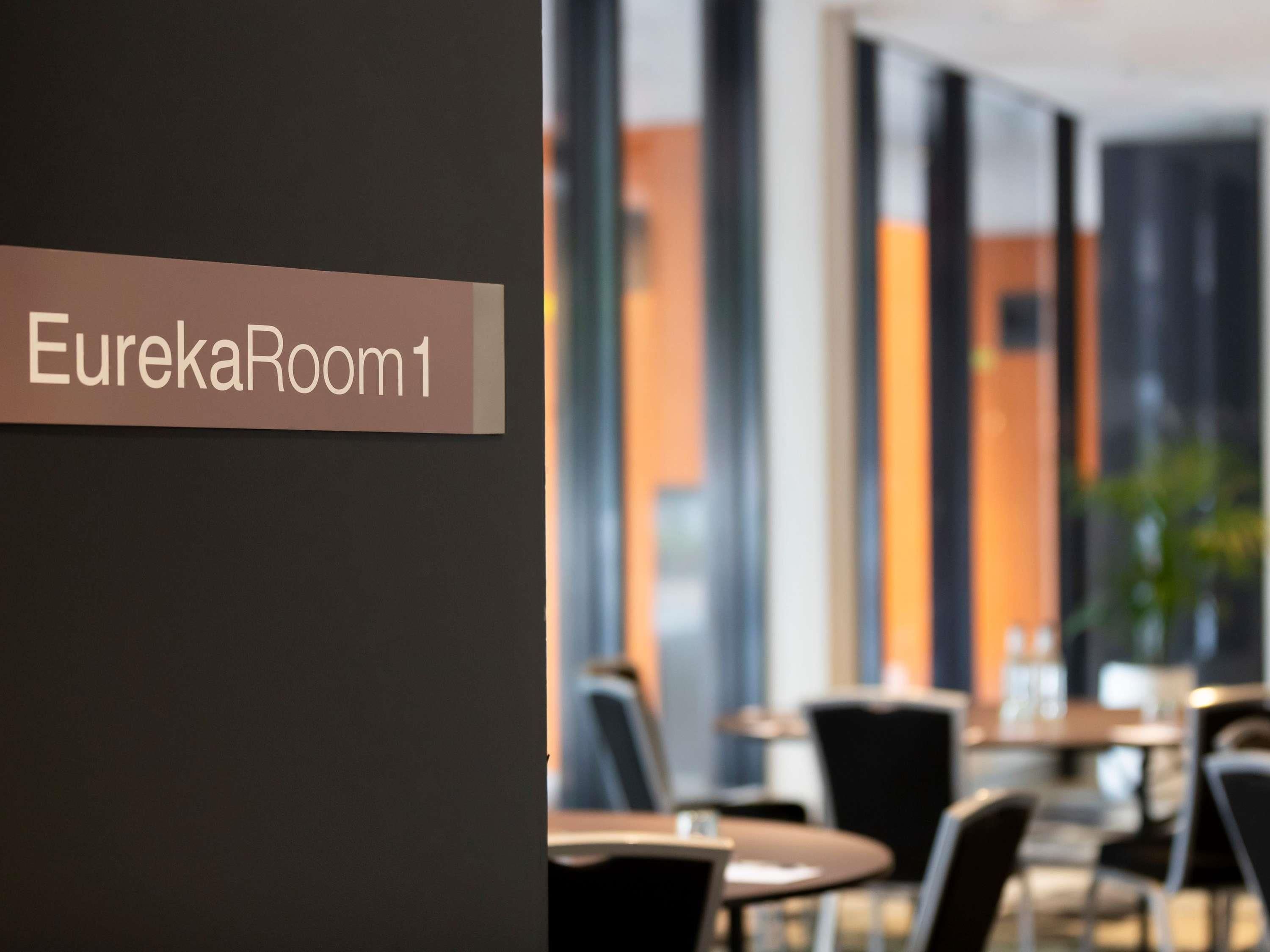 A sign reading 'EurekaRoom1' in a modern hotel lobby with large windows and a seating area.