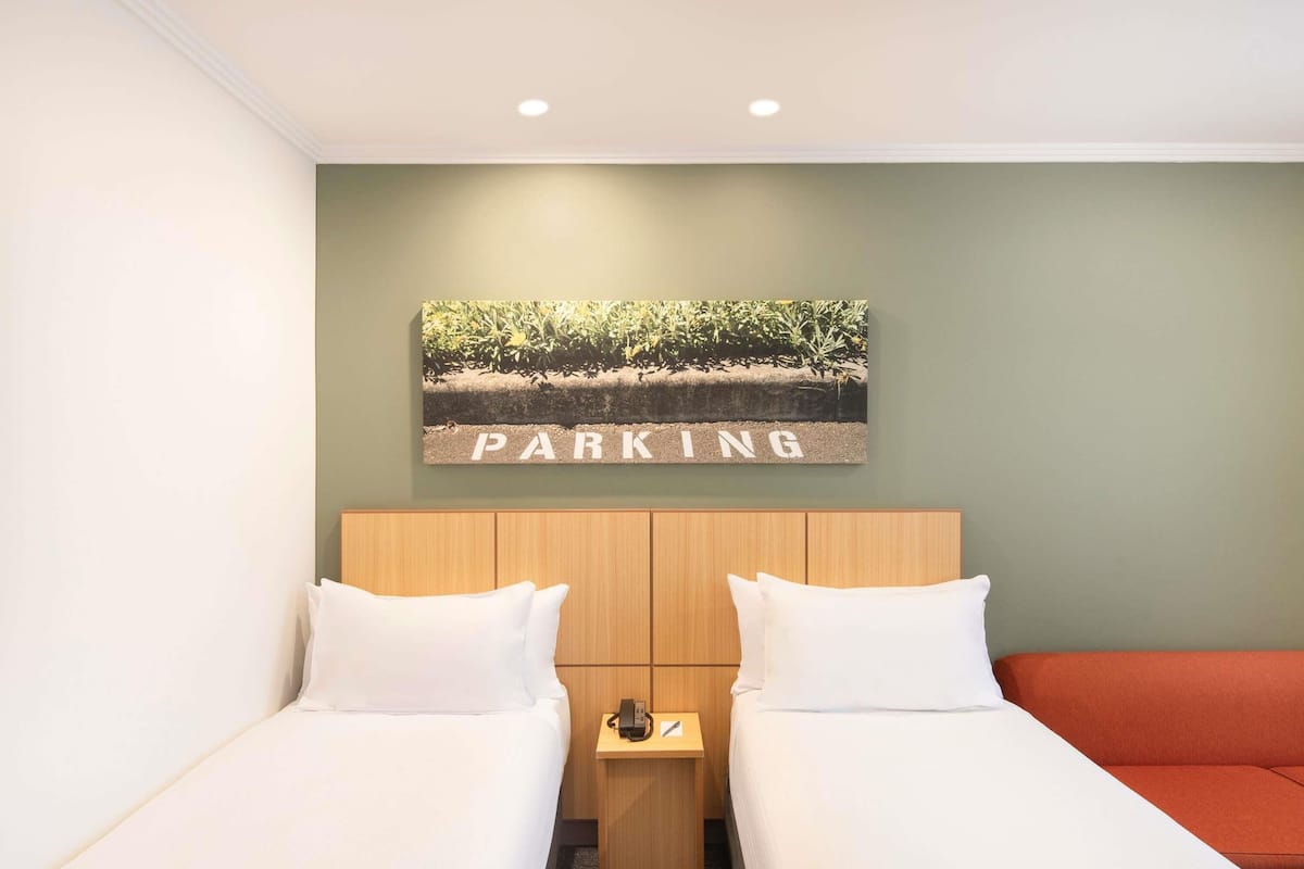 Une chambre d’hôtel avec deux lits, une petite table avec un téléphone et un tableau accroché au mur représentant un parc, avec le mot « PARKING » écrit en dessous.