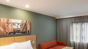 Habitación de hotel con cama, cabecera de madera, sofá rojo, mesita de madera y una ventana grande con cortinas.